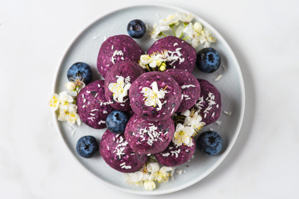 No bake blueberry and acai energy bites or balls made of nuts, coconut flakes and dates served with flowers. top view. healthy vegan food dessert